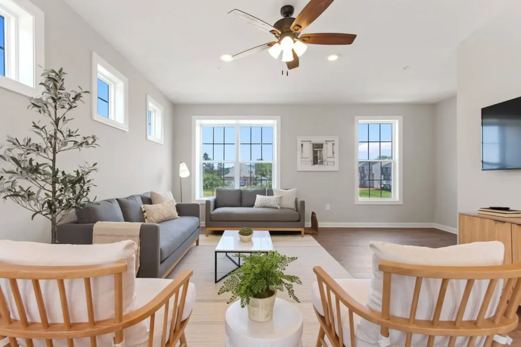 Model living room with gray sofas, wood-frame chairs, ceiling fan, and three large grid windows