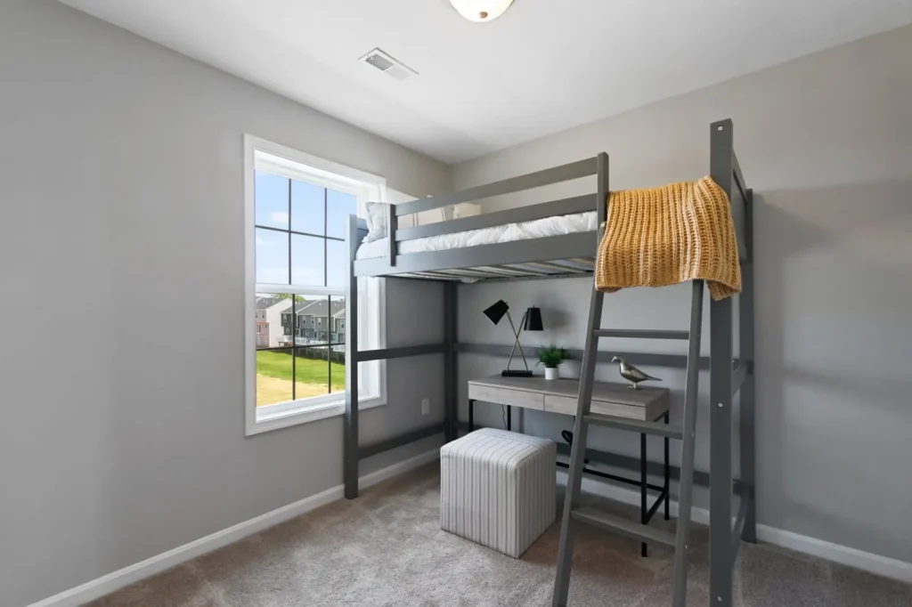 Gray loft bed with built-in desk, striped ottoman, and window view of nearby townhomes