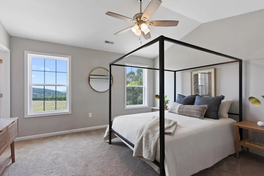 Sunlit master bedroom with black four-poster bed, neutral linens and two windows showing rolling fields